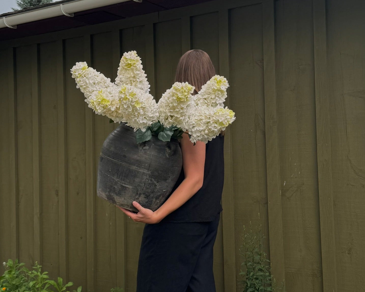 Person holding a large pot of white flowers against a green wall.